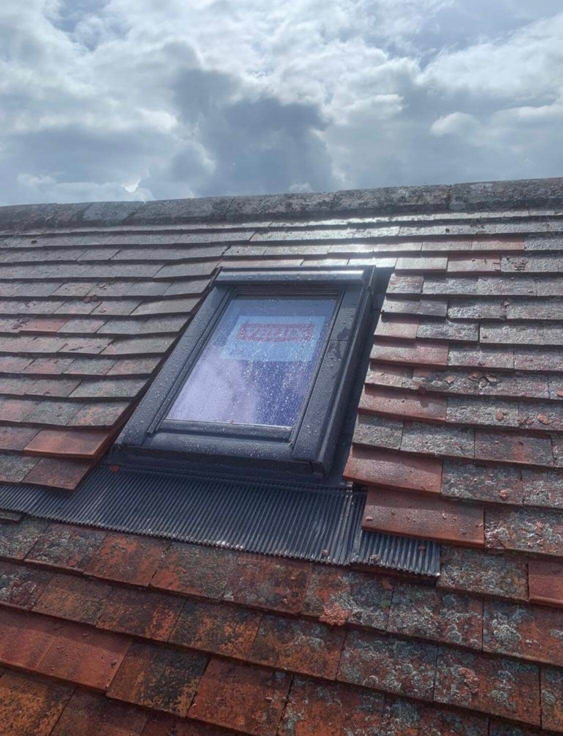 Skylight on a red-tiled roof against a cloudy sky. Black frame, protective film on glass, and black underlayment.