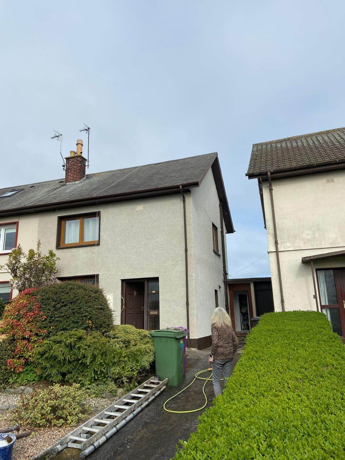 Two-story house with a person walking away. Green shrubs and a recycling bin are in front.