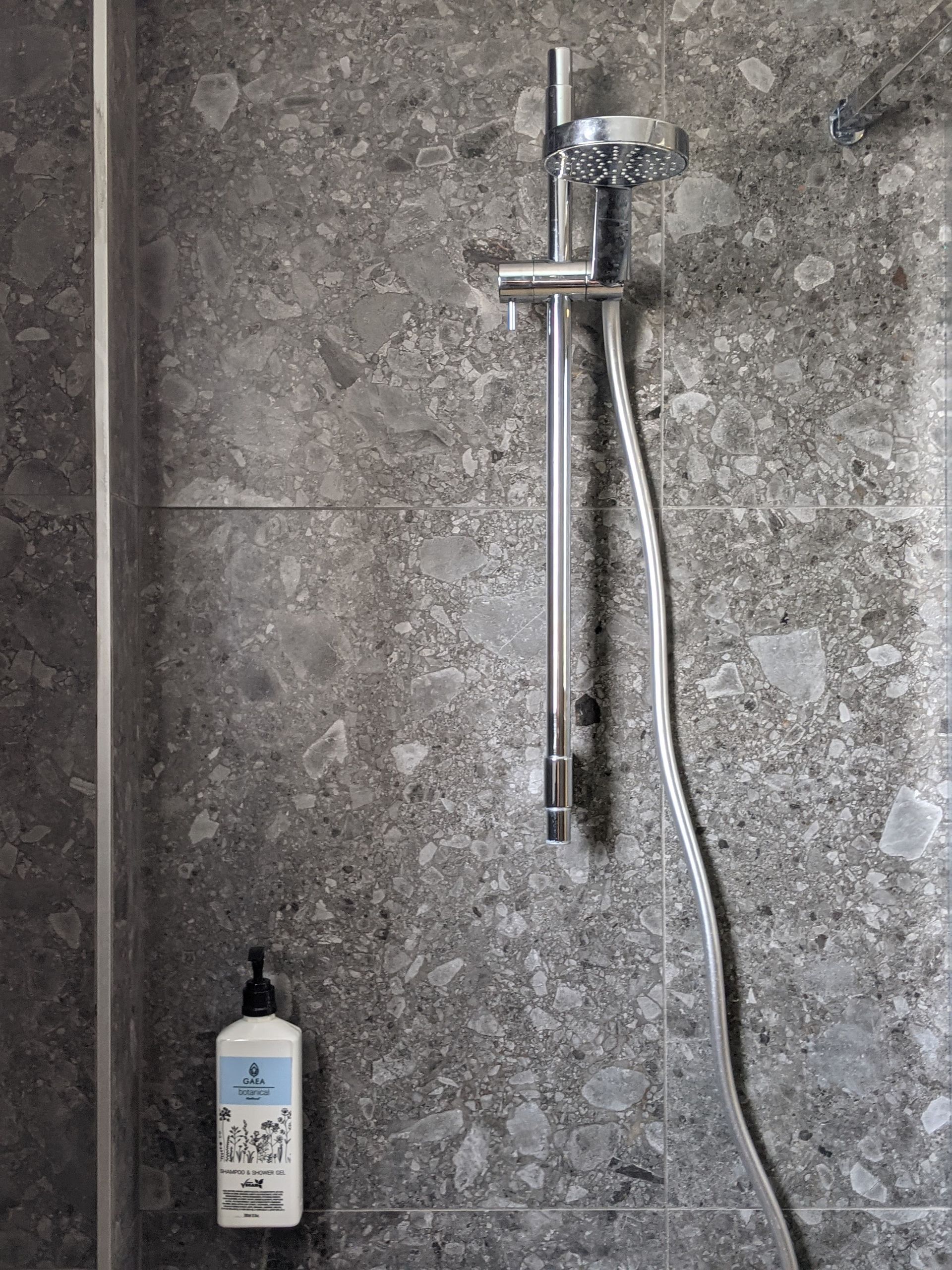 Shower with gray stone tile walls and a silver shower head, and a soap dispenser.