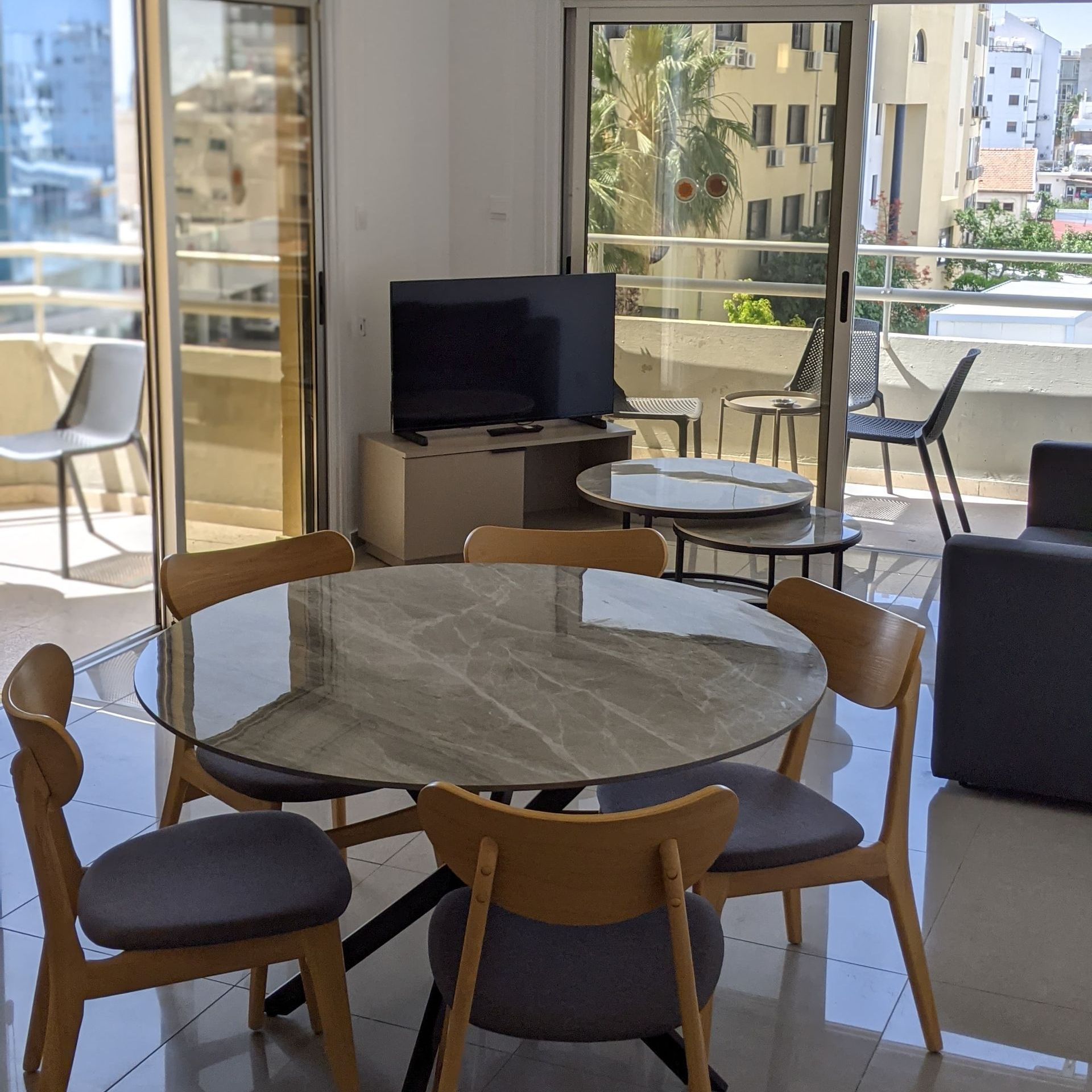 A modern dining area with a round table and six chairs, overlooking a balcony with city views.