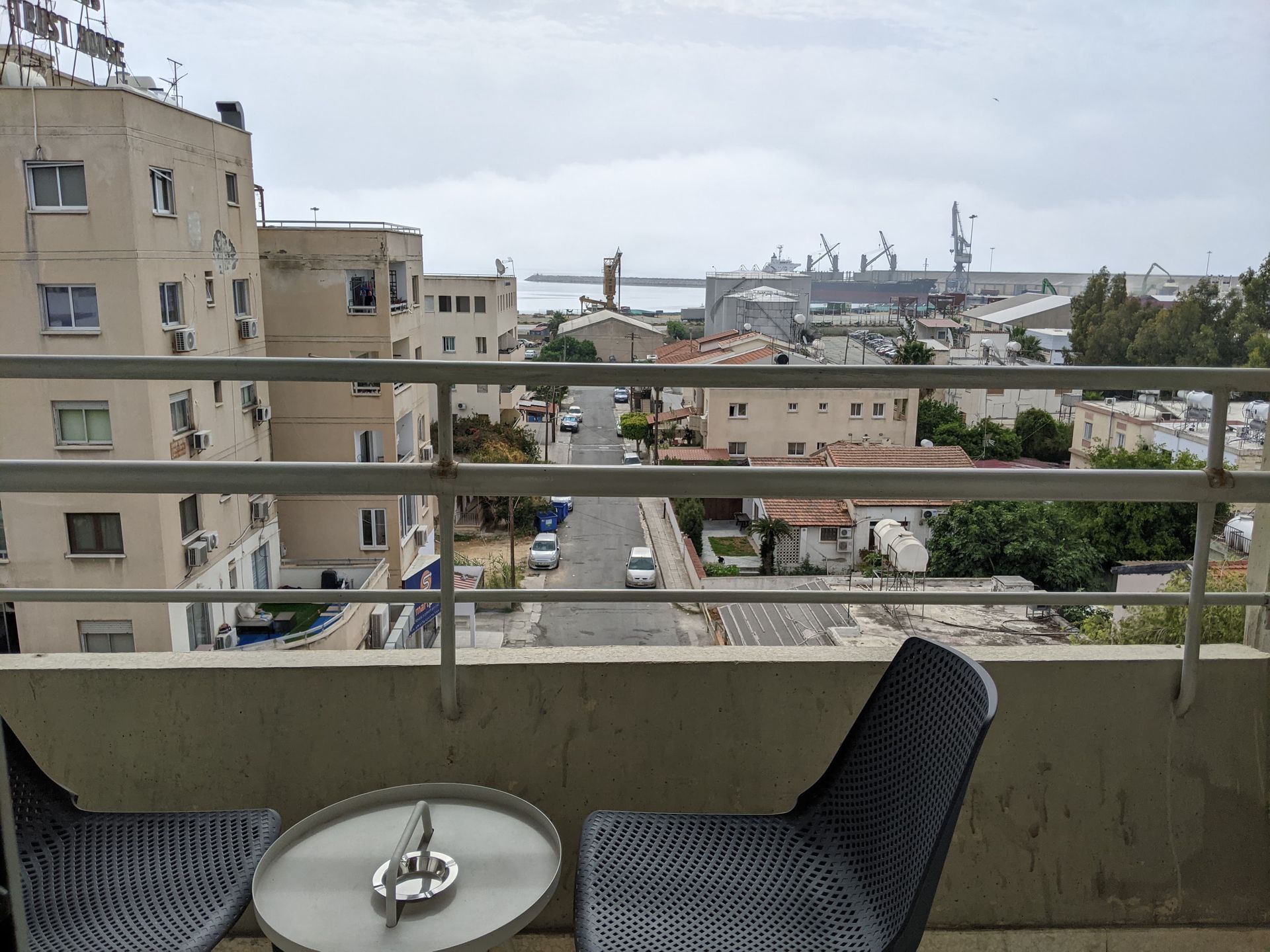 Balcony view overlooking a street and port. Two chairs and a small table in the foreground. Buildings, a road, and harbor cranes in the distance. Overcast sky.