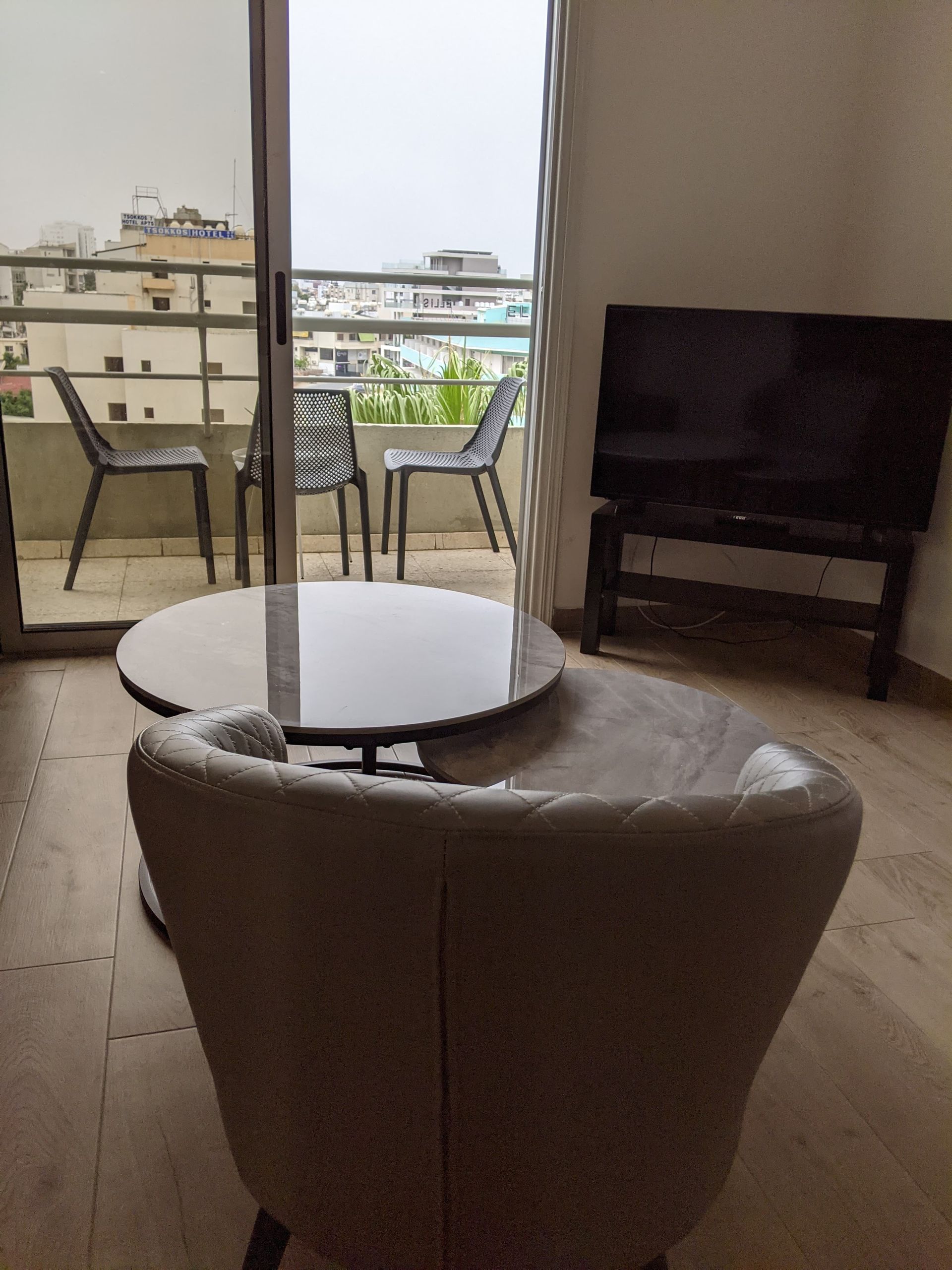 Interior view of a modern apartment with a balcony. Includes a round table with a chair in the foreground and a TV on a stand.