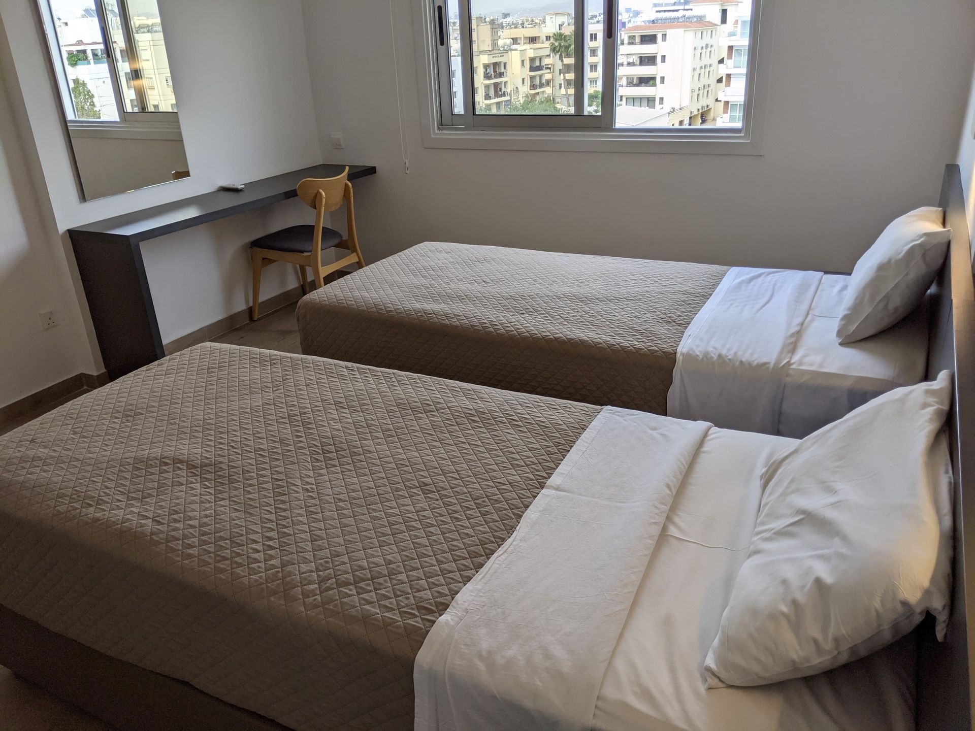 Two twin beds in a bright room with a desk and window overlooking a city. Light brown bedding, white sheets.