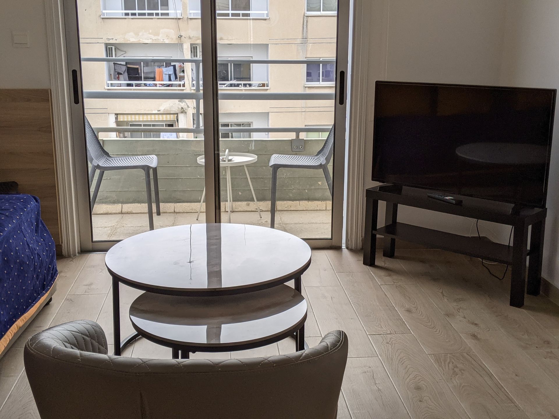 A small living space with a balcony. Includes a round coffee table, a TV on a stand, and a chair.