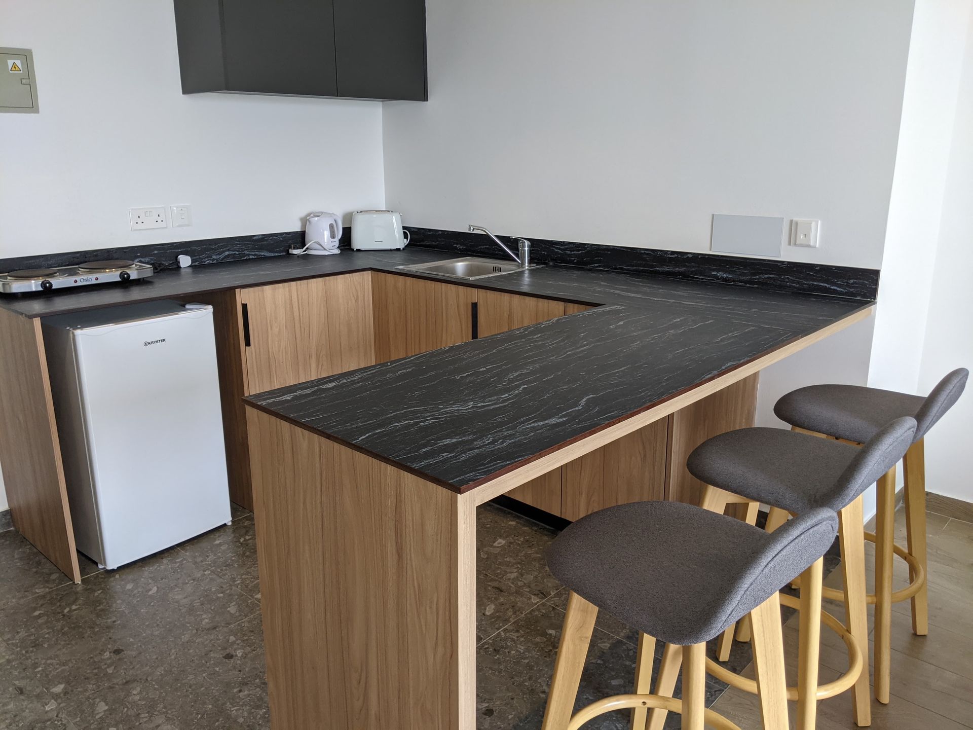 Small modern kitchenette with a countertop, cabinets, and three bar stools. Includes a mini-fridge, sink, and microwave.