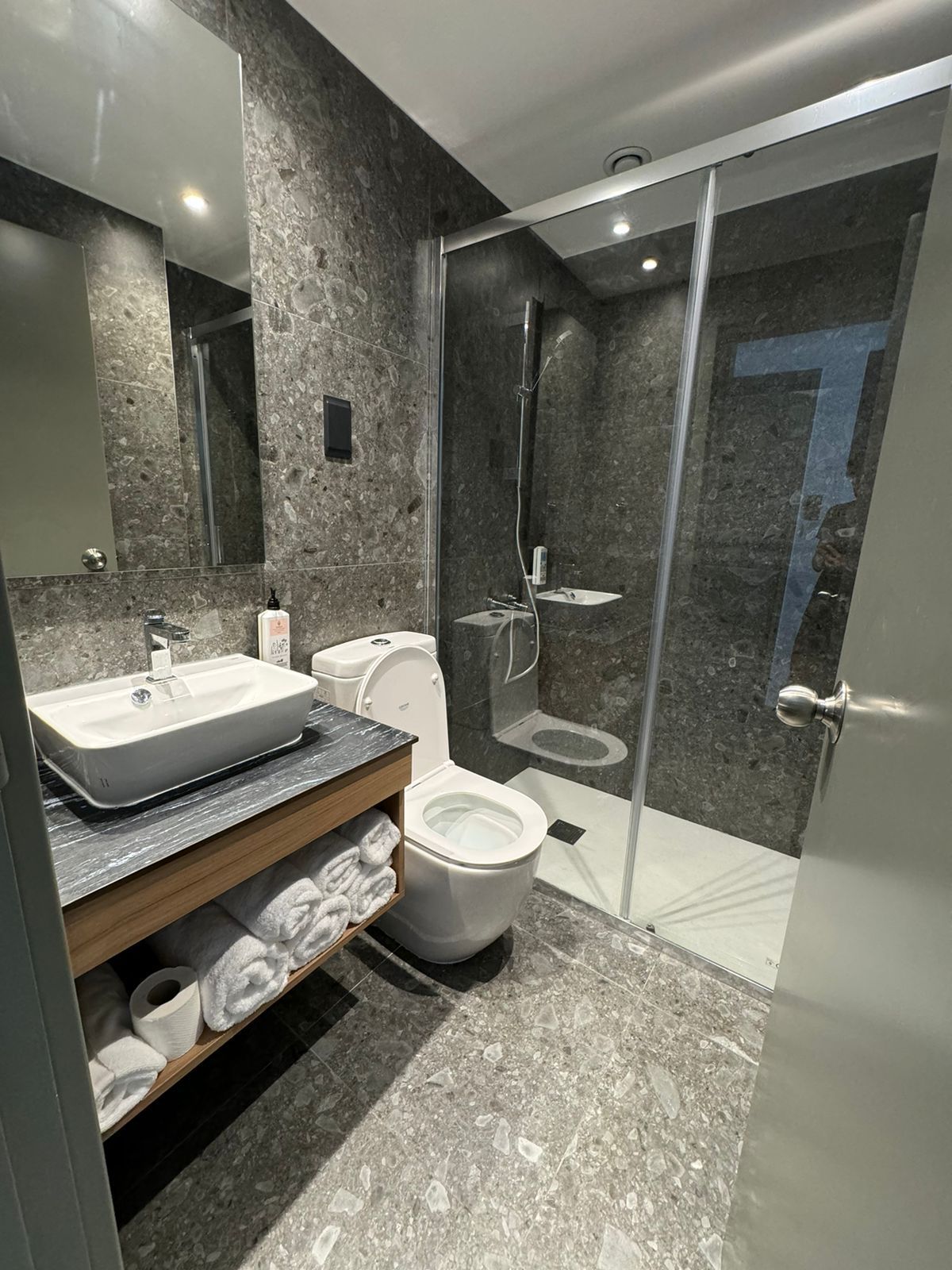 Modern bathroom with gray tile, featuring a sink, toilet, and glass shower. White towels are stacked in an open shelf.