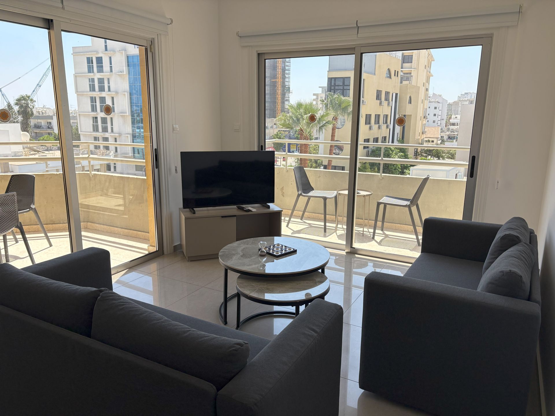Living room with a balcony view, featuring a grey armchair, nested coffee tables, and a TV. The balcony overlooks a cityscape with the sea.