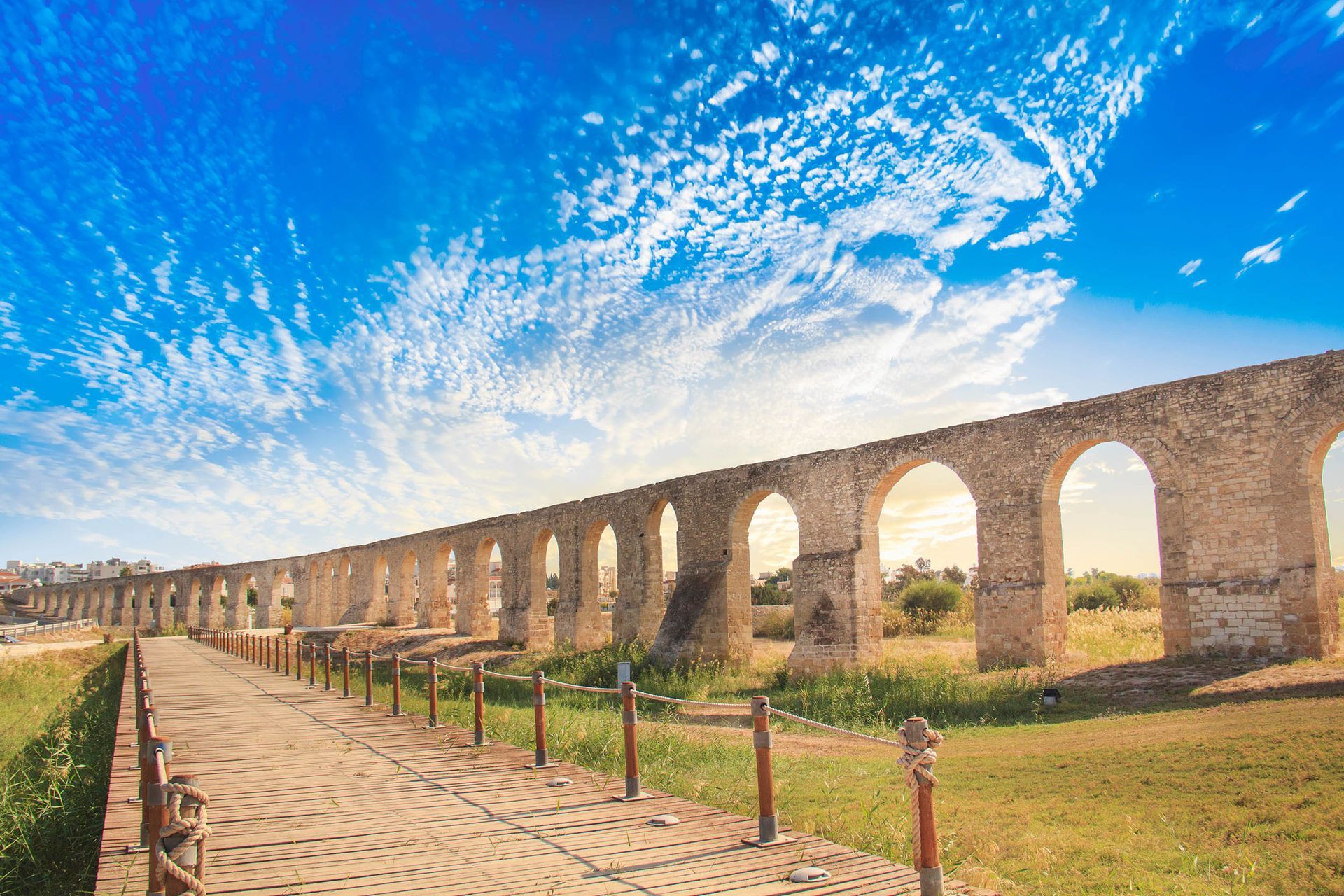 kamares aqueduct A long stone aqueduct surrounded by grass and trees under a blue sky with clouds.