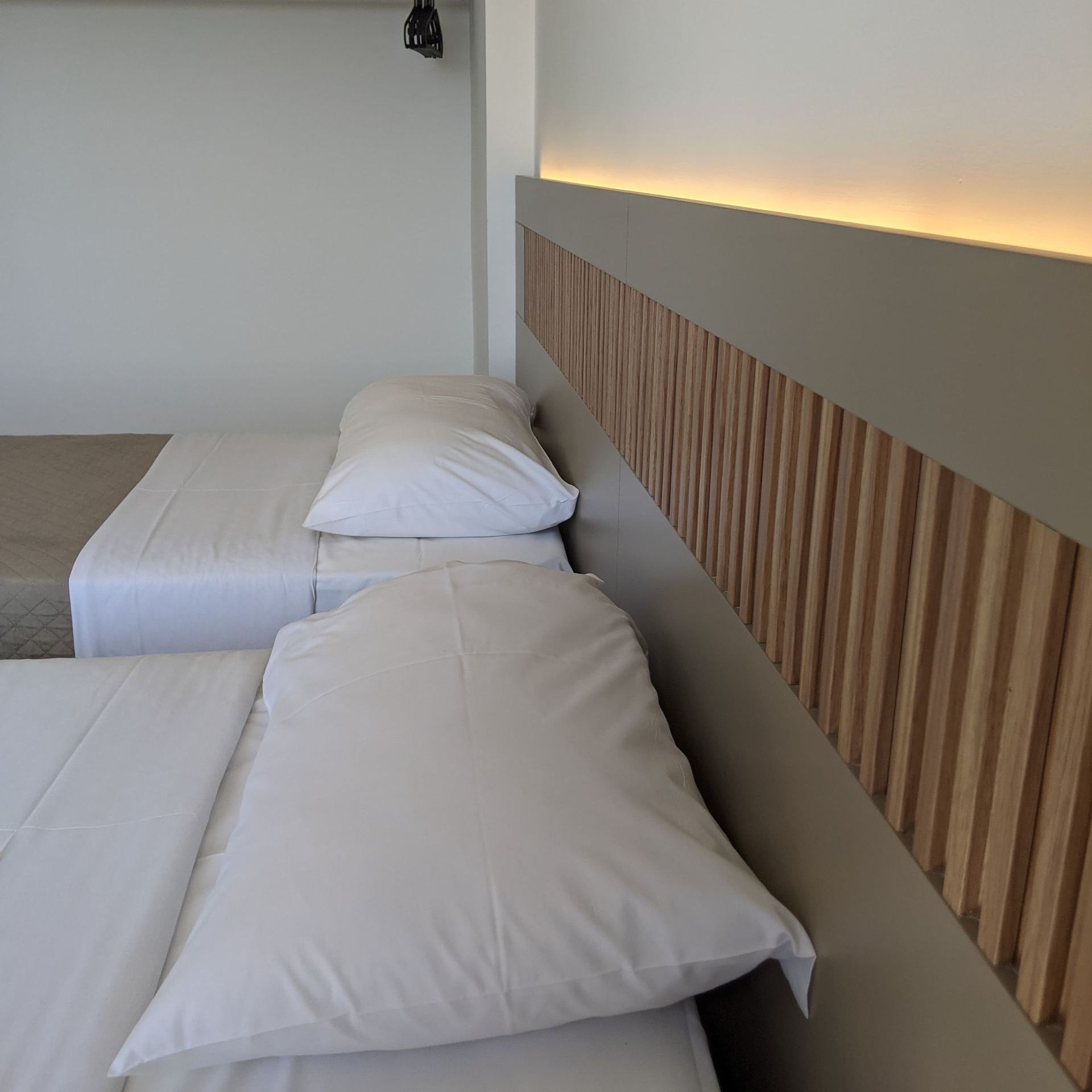 Two twin beds with white linens and pillows. A headboard with wood panels is illuminated from above.