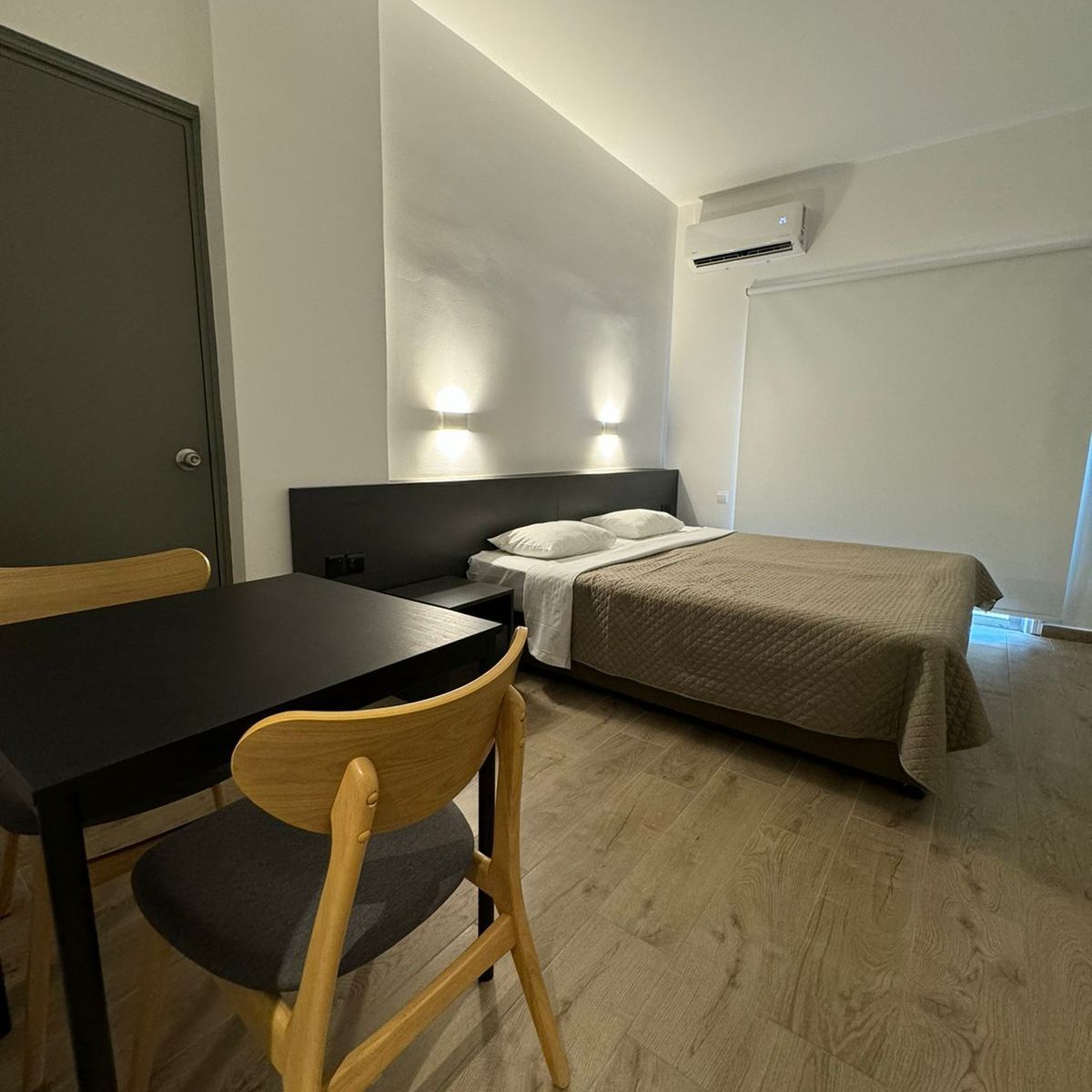 A modern studio apartment with a bed, small table with chairs, and a closed gray door.