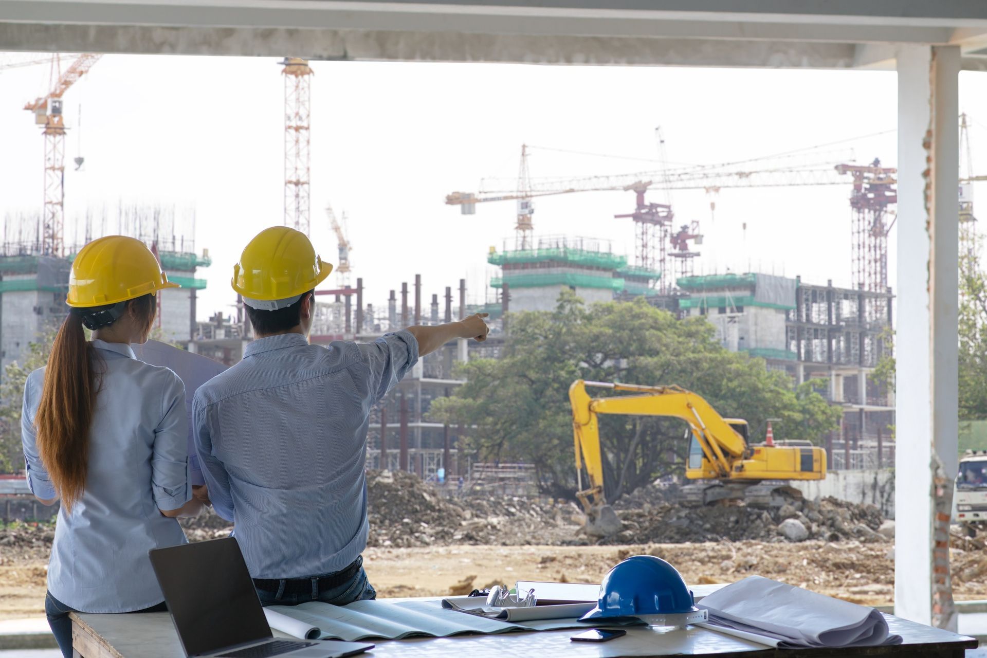 Two people in hard hats look at a construction site with cranes, pointing at a building.