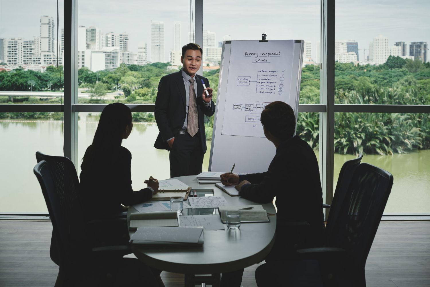 Man presenting at whiteboard to colleagues in a modern office with city views.