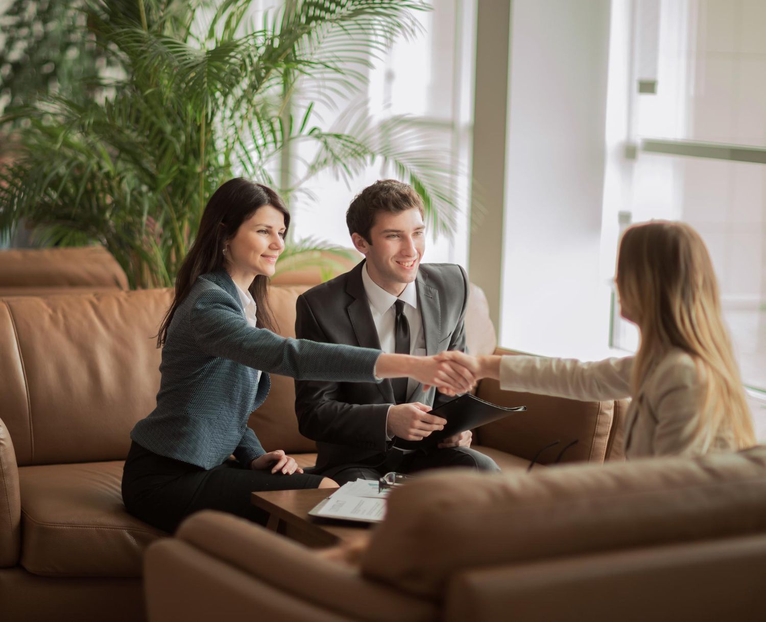 People in business attire shaking hands in a bright office setting; two on a brown sofa, one reaching out.