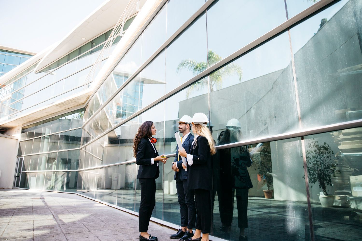 Group of professionals in suits and hard hats discuss plans in front of a modern glass building.