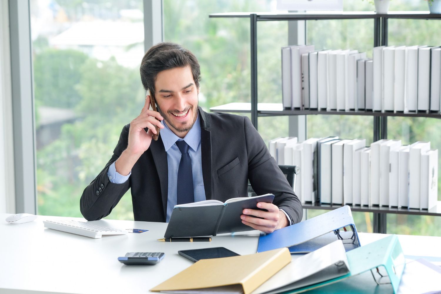 Man in suit smiles while on phone, looking at notebook in bright office.