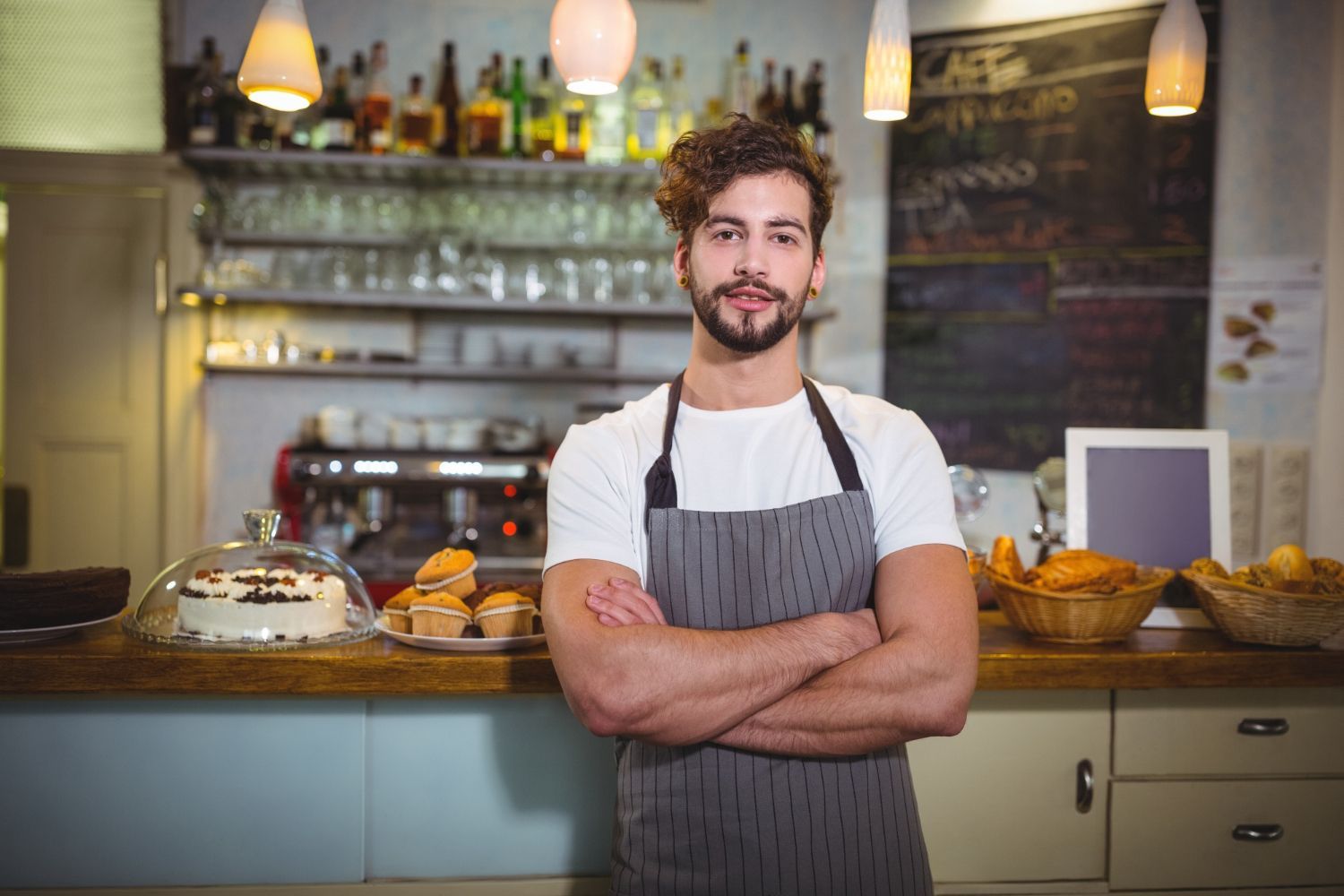 Man in apron at cafe counter with arms crossed, smiling.