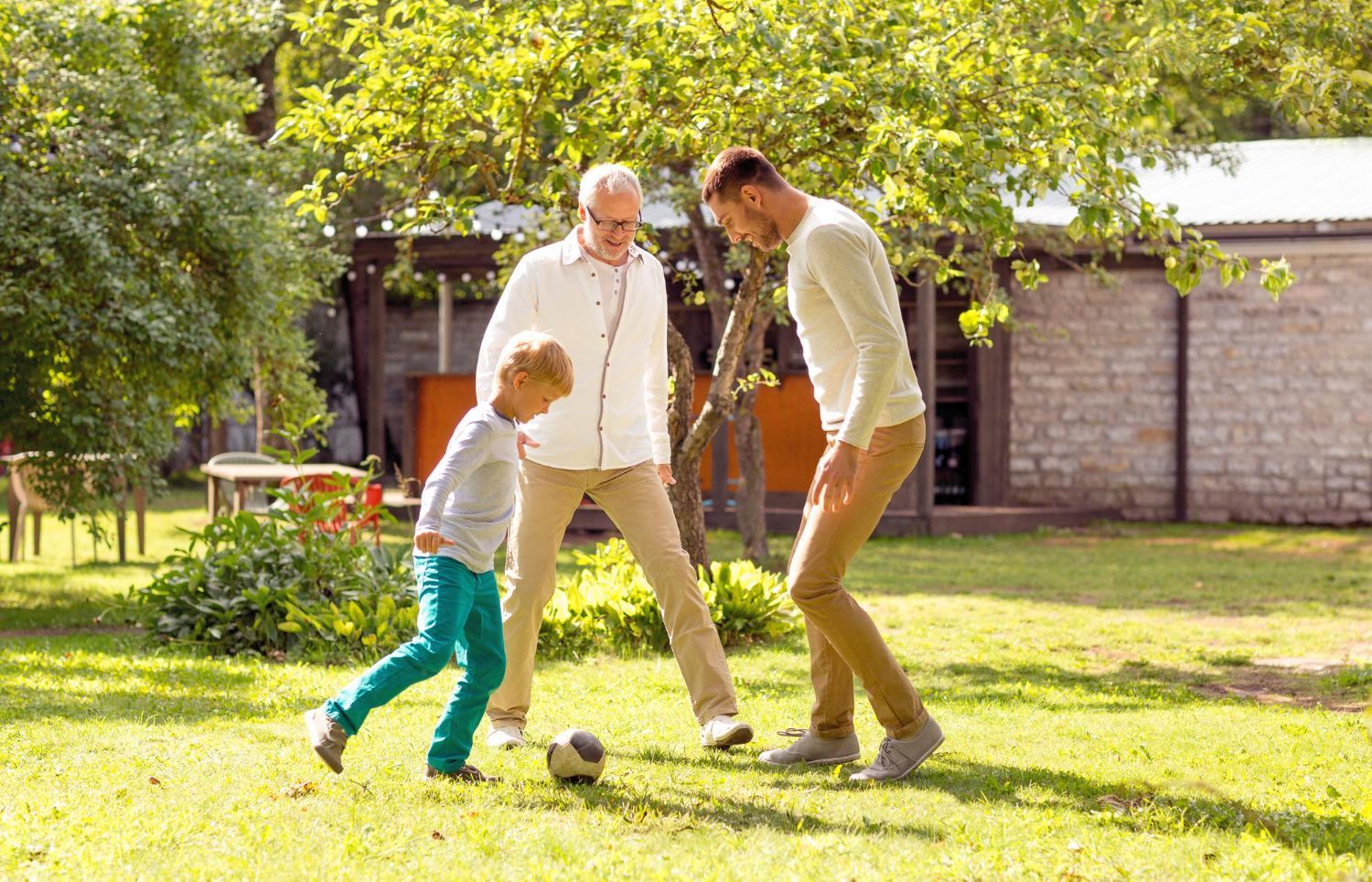 A young boy, man, and older man play soccer on a sunny lawn.