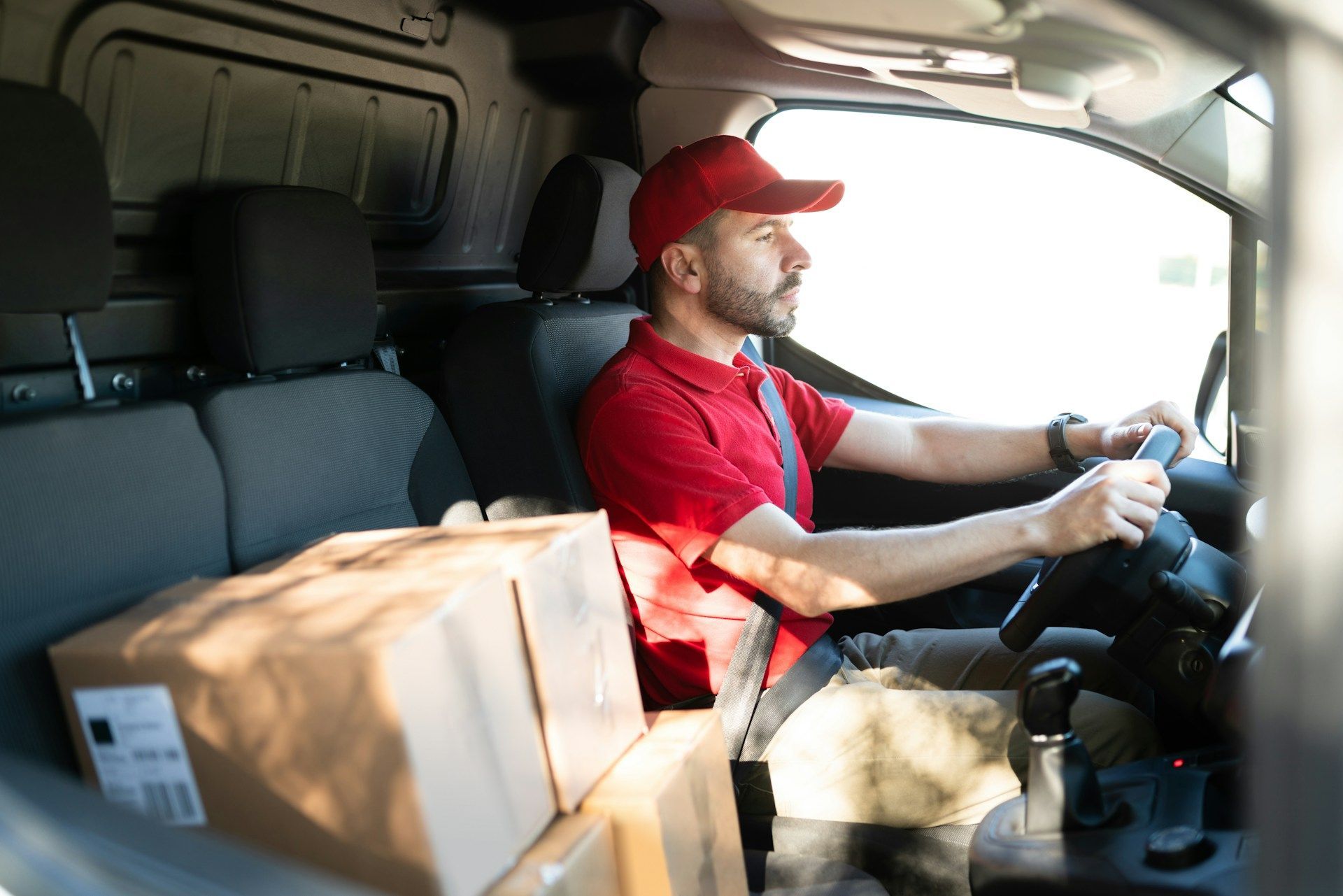 Delivery driver in red shirt and cap drives van, boxes in the seat.