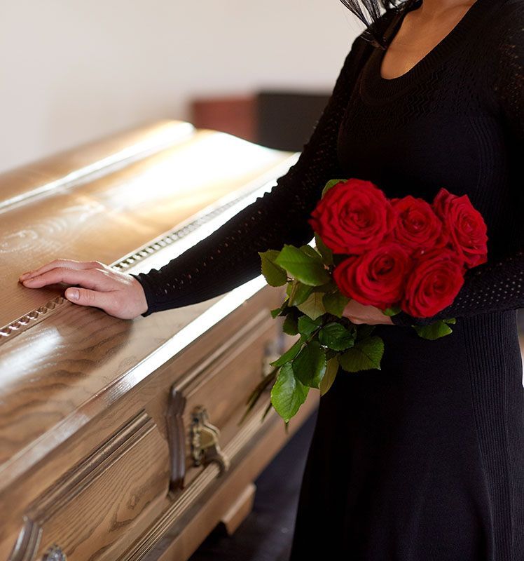 Person in black dress holding a bouquet of red roses beside a wooden coffin