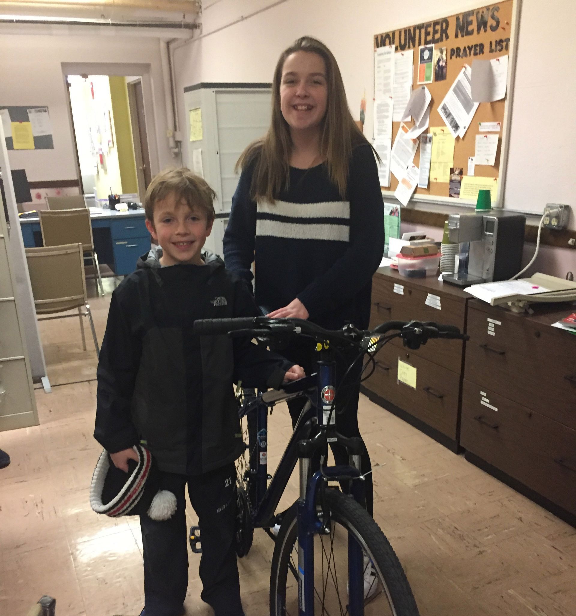 A girl and a boy standing next to a bicycle in front of a bulletin board that says volunteer news