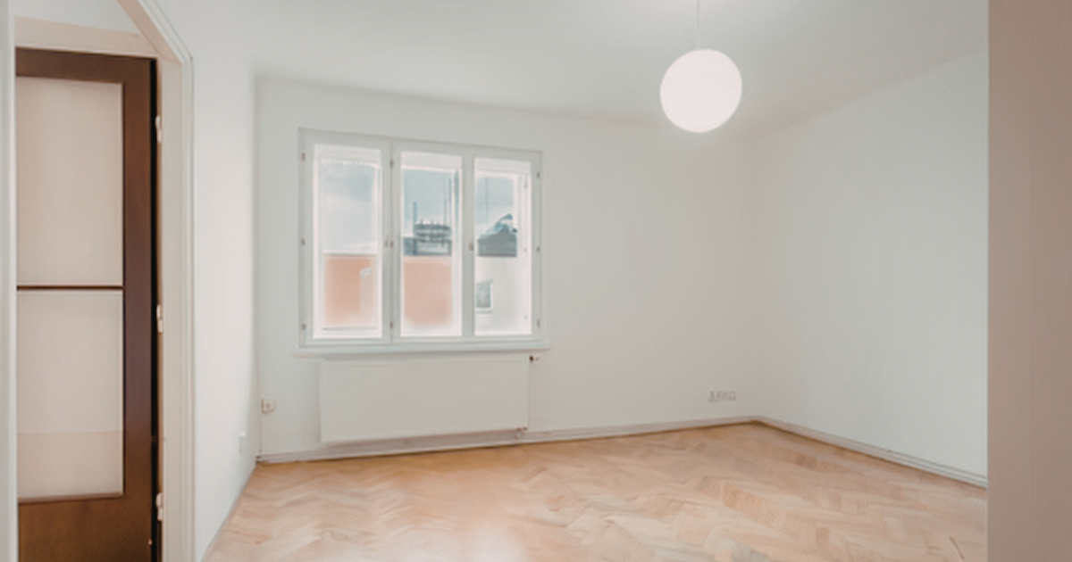 An empty room with white walls, a single window, a wall-mounted radiator, and light-colored herringbone wood floors.