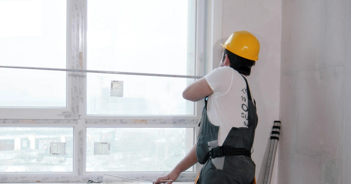 A person in a yellow hard hat and work overalls measuring the frame of a large window with a tape measure.