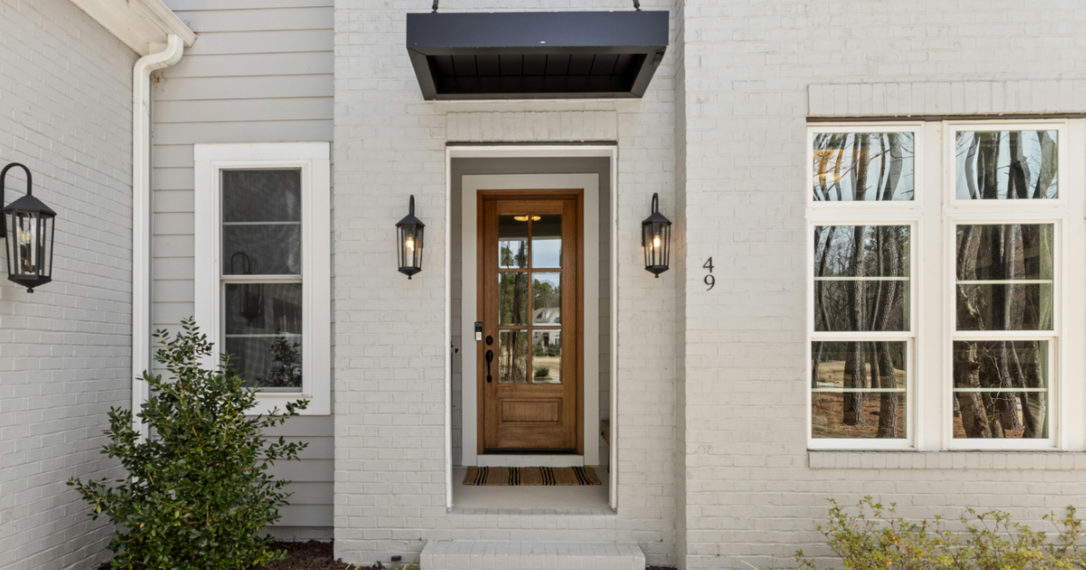 A light gray brick house exterior with a wooden front door, black light fixtures, and a modern black metal awning.