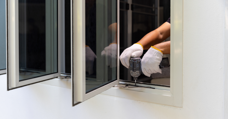 A pair of hands in white work gloves uses a power drill to install or repair a white-framed glass window.