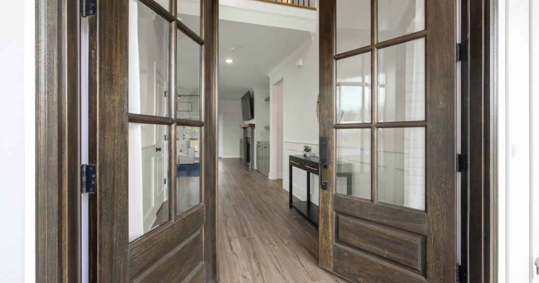 Double dark wood French doors with glass panes open into a hallway with hardwood floors and a console table.