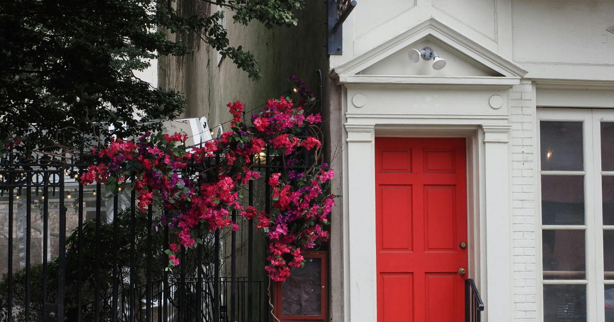A bright red front door with a white classical frame, next to a black iron fence adorned with deep pink climbing flowers.