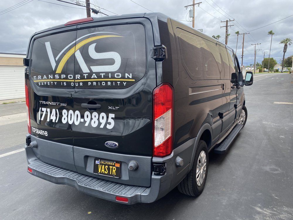 Black VKS Transportation van with logo and phone number, parked on a street.