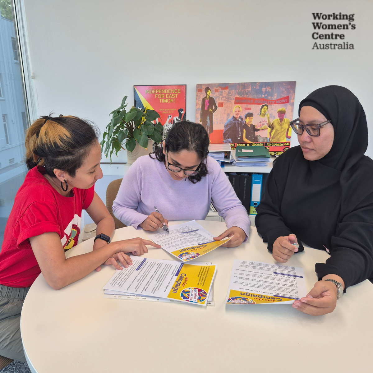 Three women sitting at a round table in the WWCA office, reviewing printed NDA campaign materials.