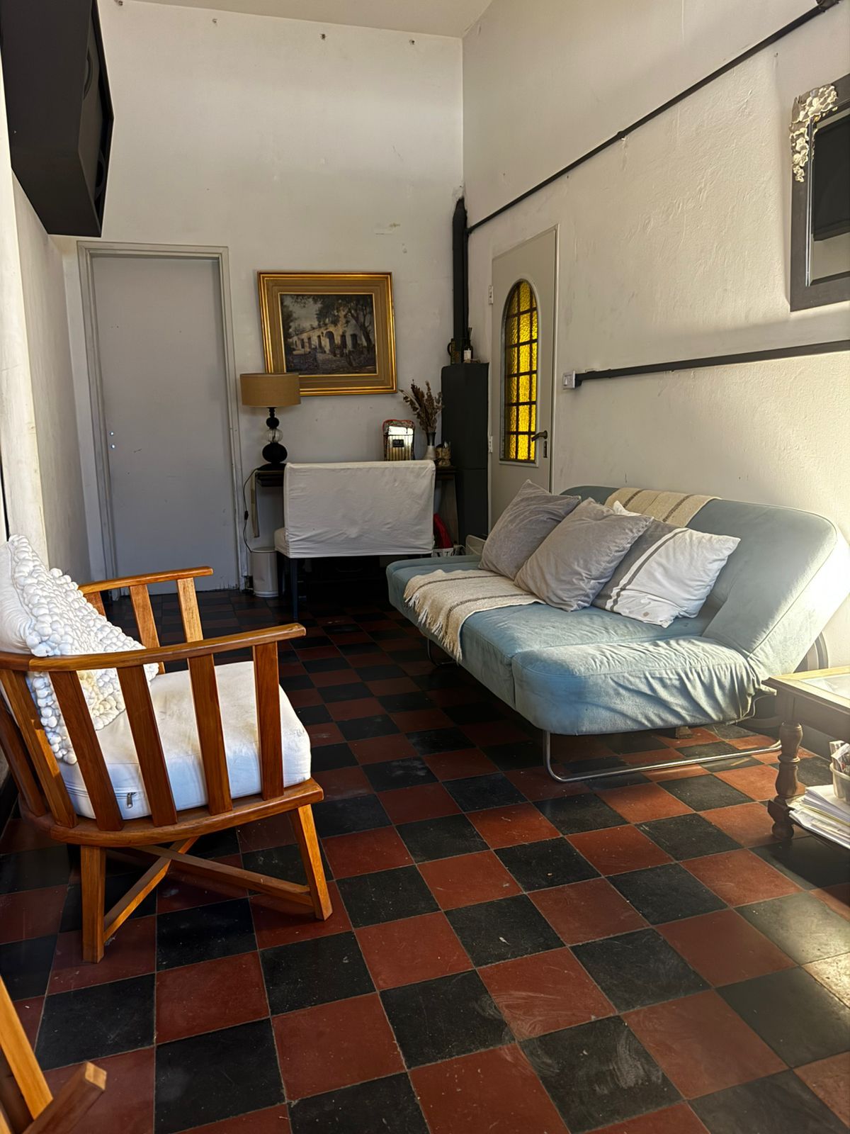 Una sala de estar con un futón azul, una silla de madera con cojines blancos y un suelo de baldosas a cuadros rojas y negras.