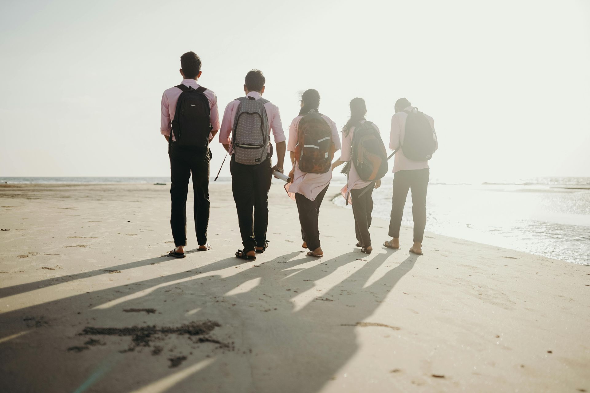 Cinco personas con mochilas caminan por una playa hacia el sol.