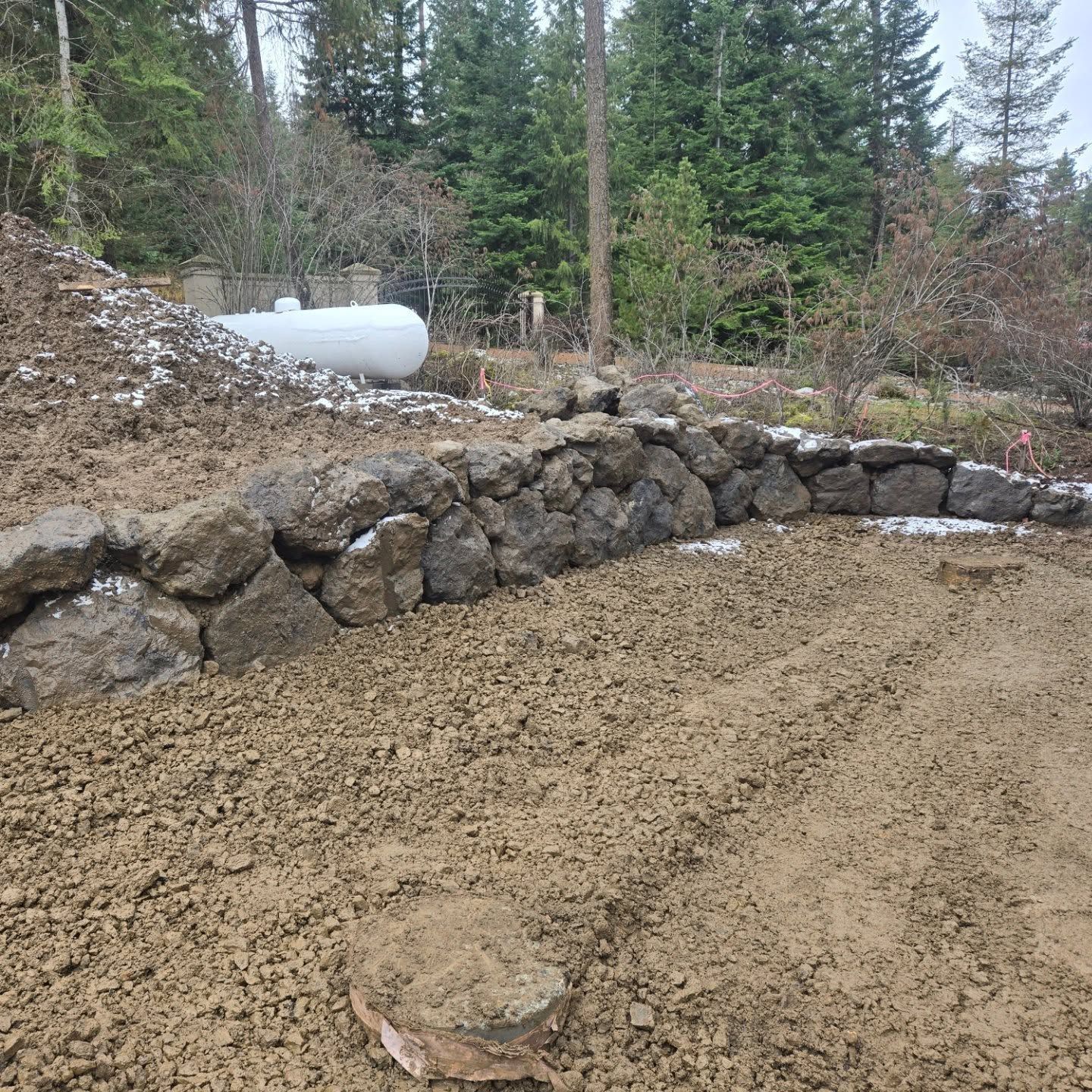 Stone retaining wall in a dirt area with a propane tank and forest background.