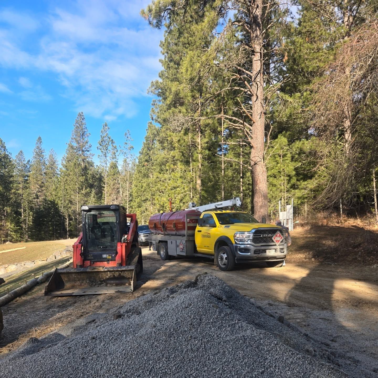 Yellow truck with a tank and small red tractor on a gravel road in a forest on a sunny day.
