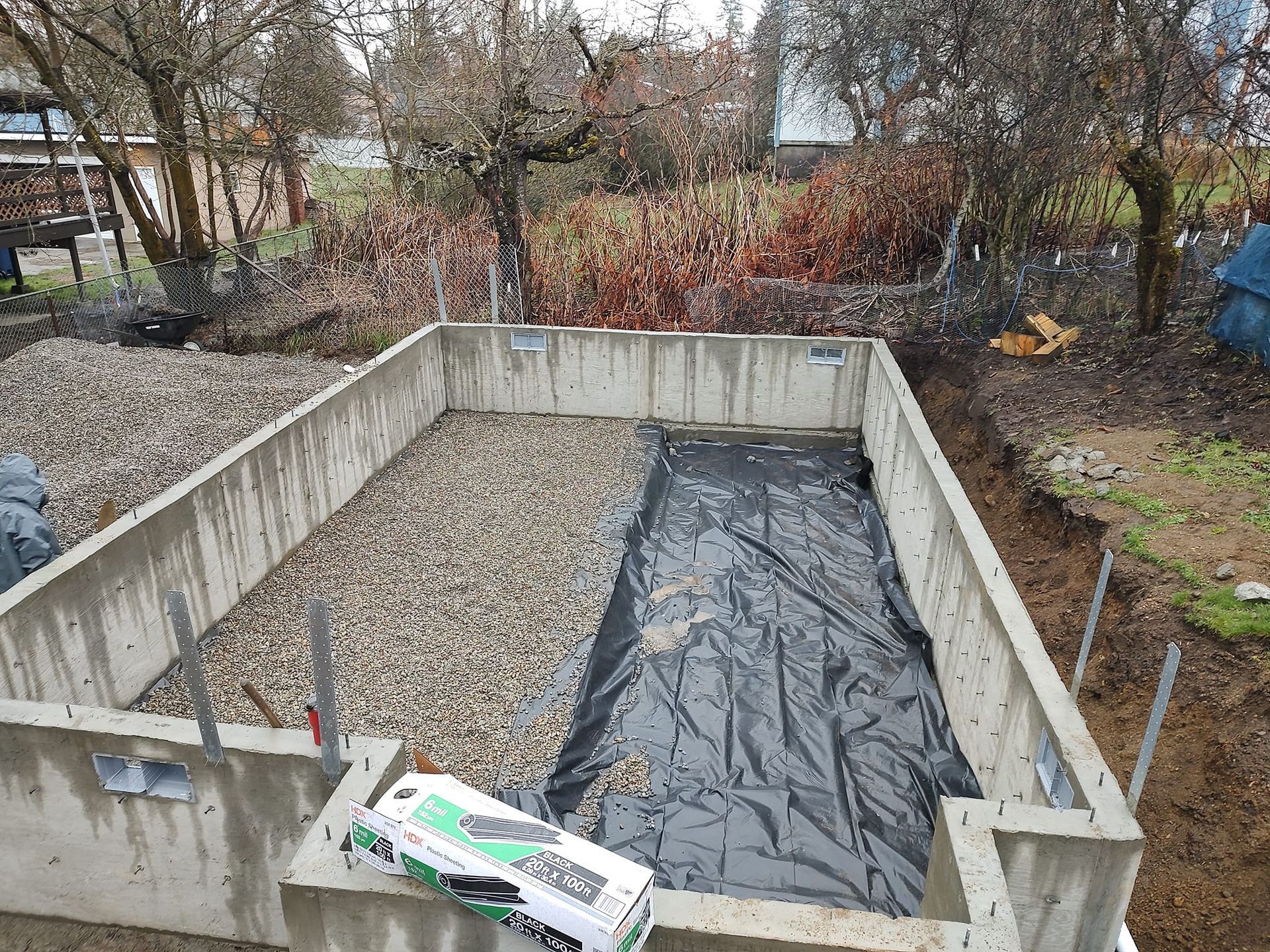 Concrete foundation with gravel, black tarp, toy truck, and surrounding yard.