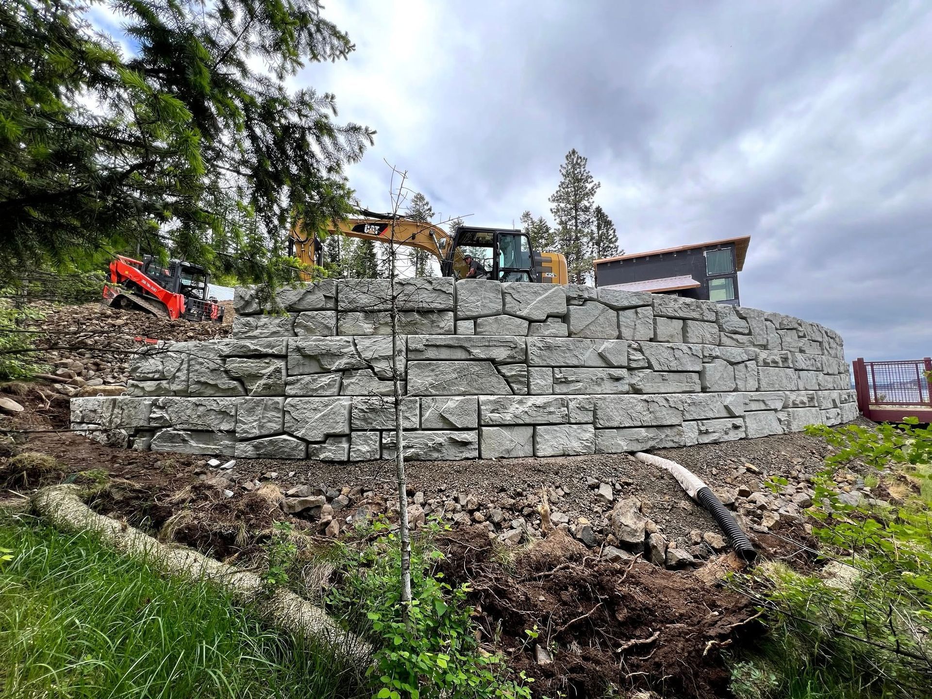 Stone retaining wall being built near a building with heavy machinery on a cloudy day.
