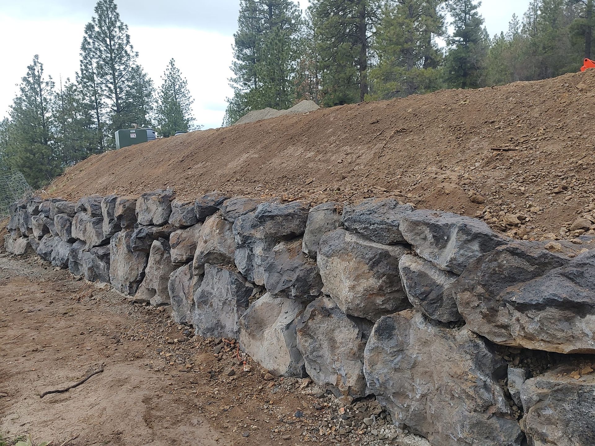 Stone retaining wall supporting a hillside of dirt, with trees in the background.