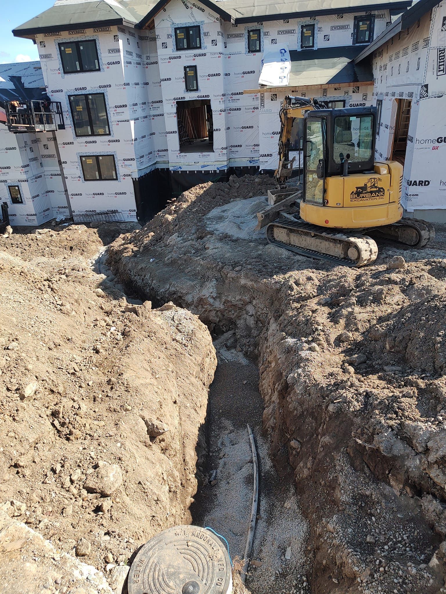 A yellow excavator digs a trench next to a building wrapped in white paper; construction site.