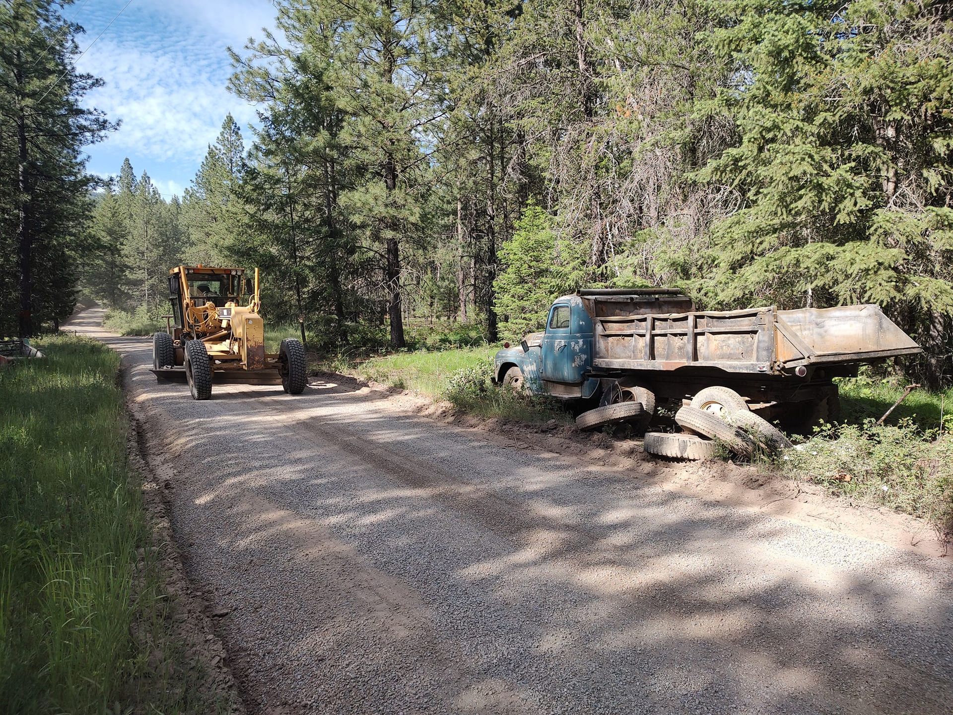 A grader and a rusted truck on a gravel road in a wooded area.