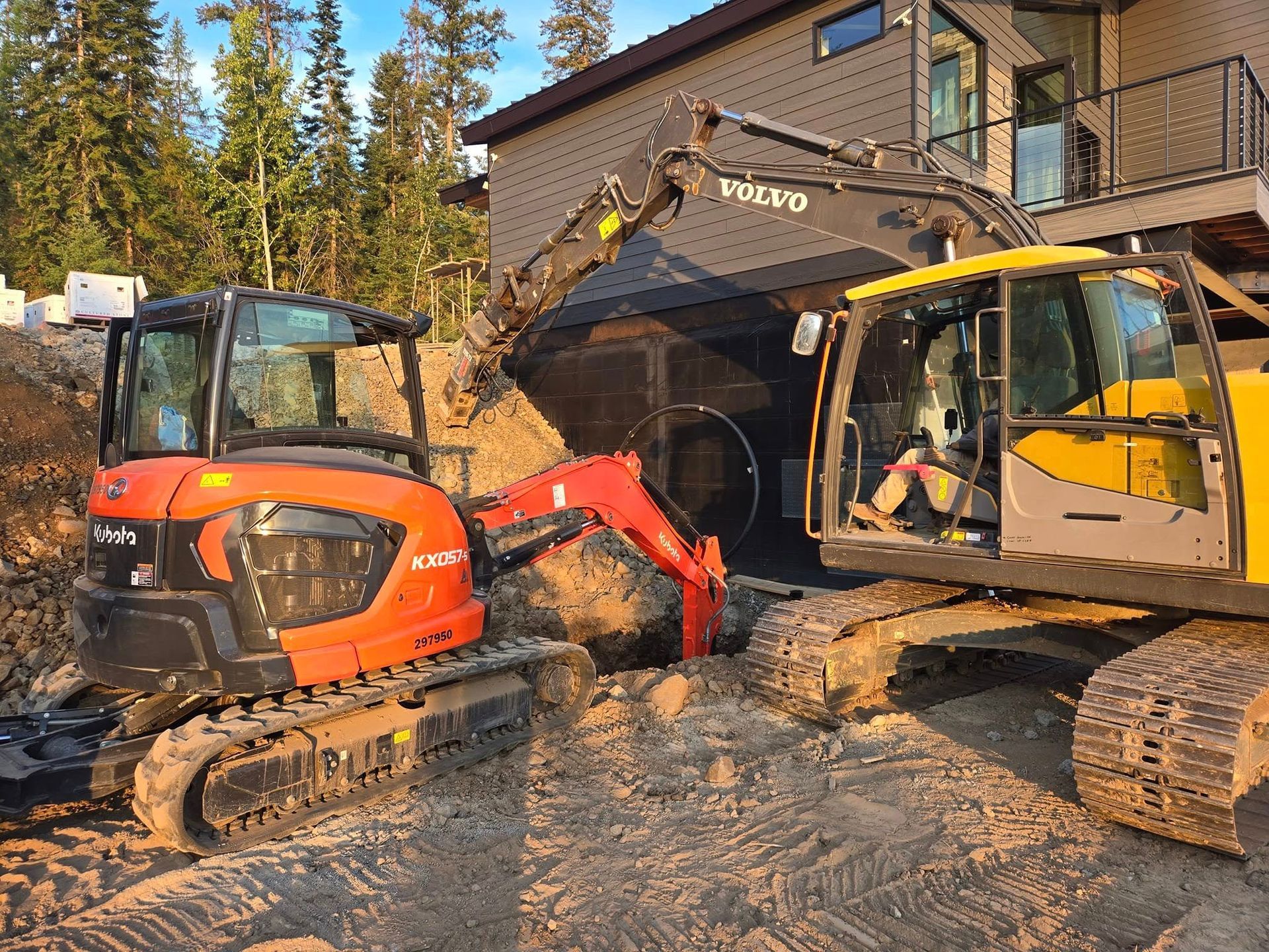 Two excavators digging dirt near a building. One is orange, the other is yellow.