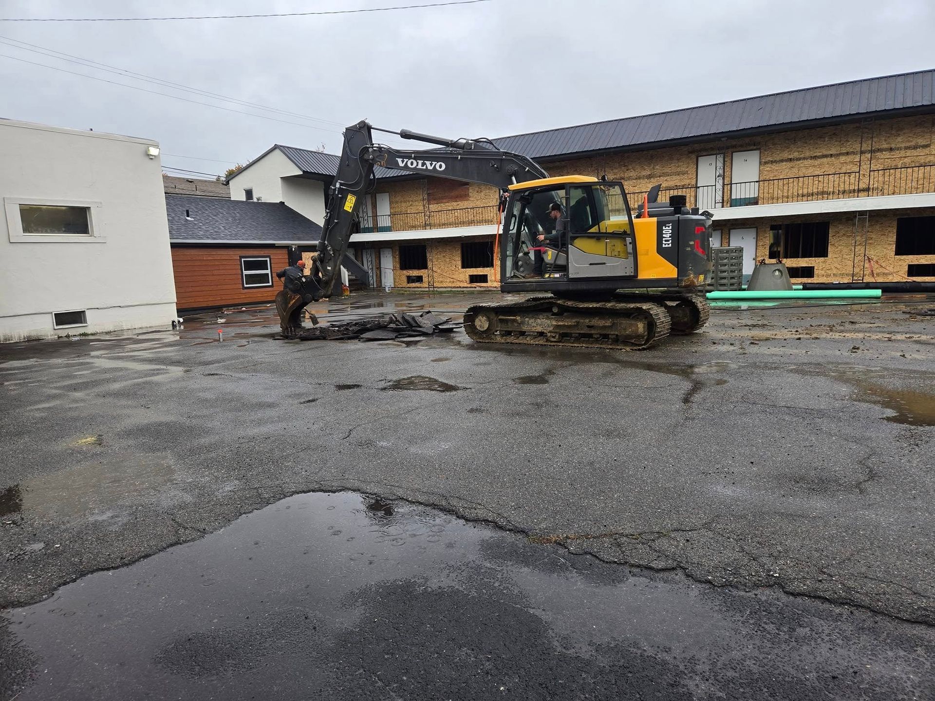 Excavator on a wet asphalt lot next to buildings under construction. Cloudy day.
