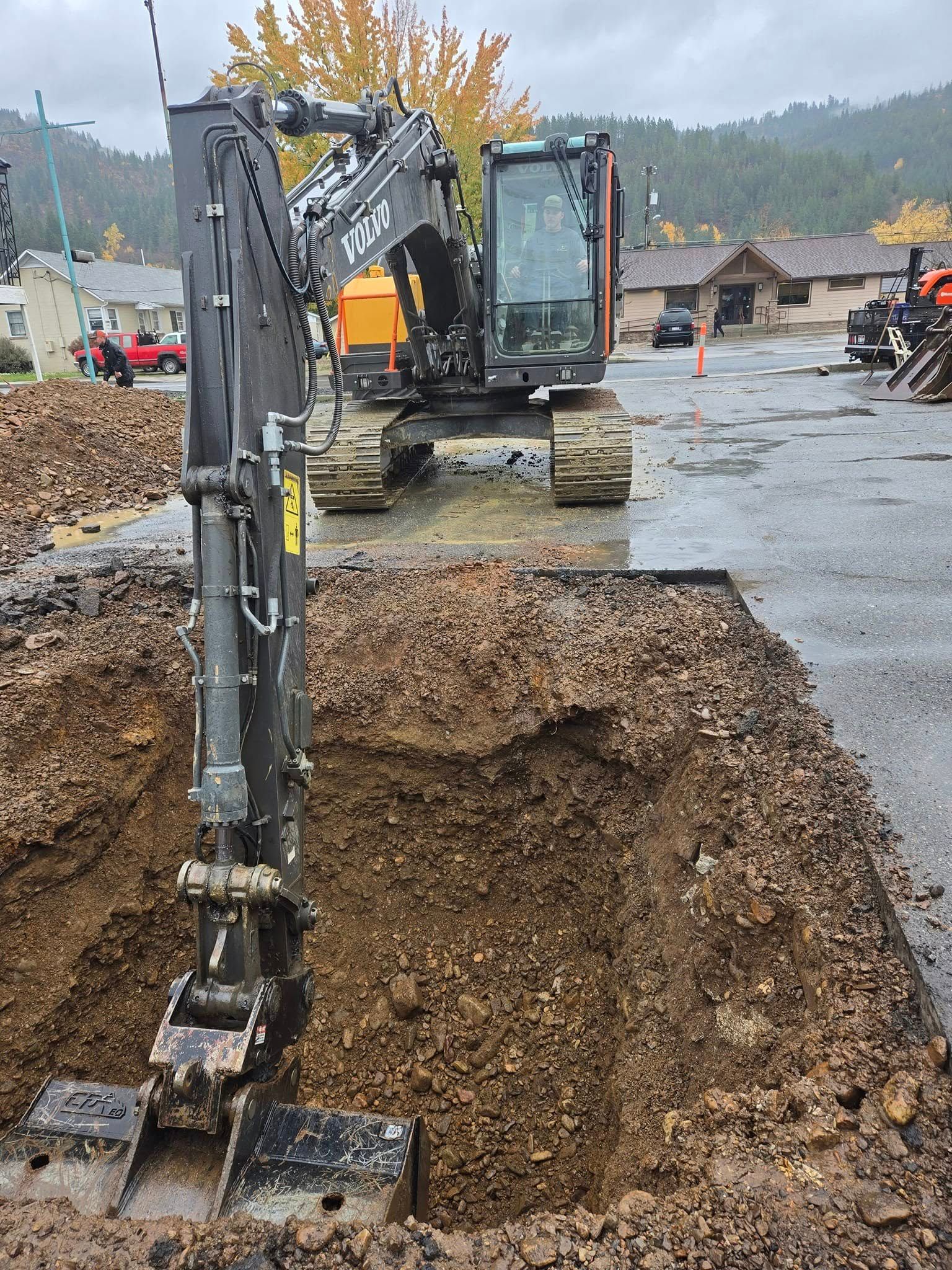 Excavator digging in a large hole in a paved area with buildings in the background.