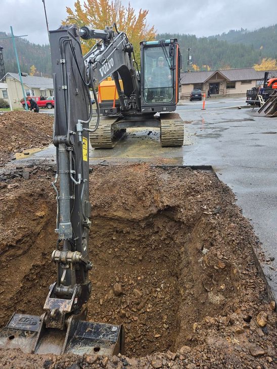 An excavator digging in a muddy pit on a street. The vehicle is black. Houses and trees are in the background.