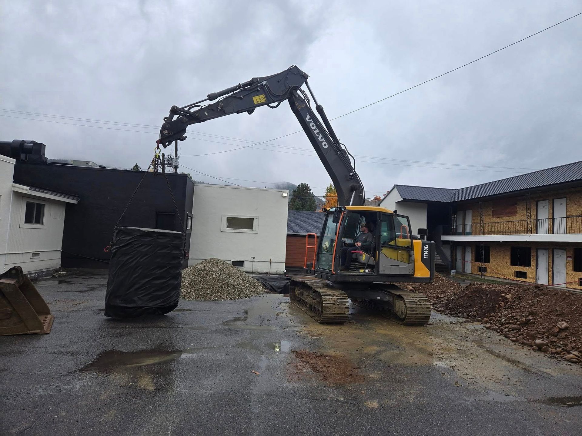 An excavator lifts a large, dark object near a building under construction on a cloudy day.