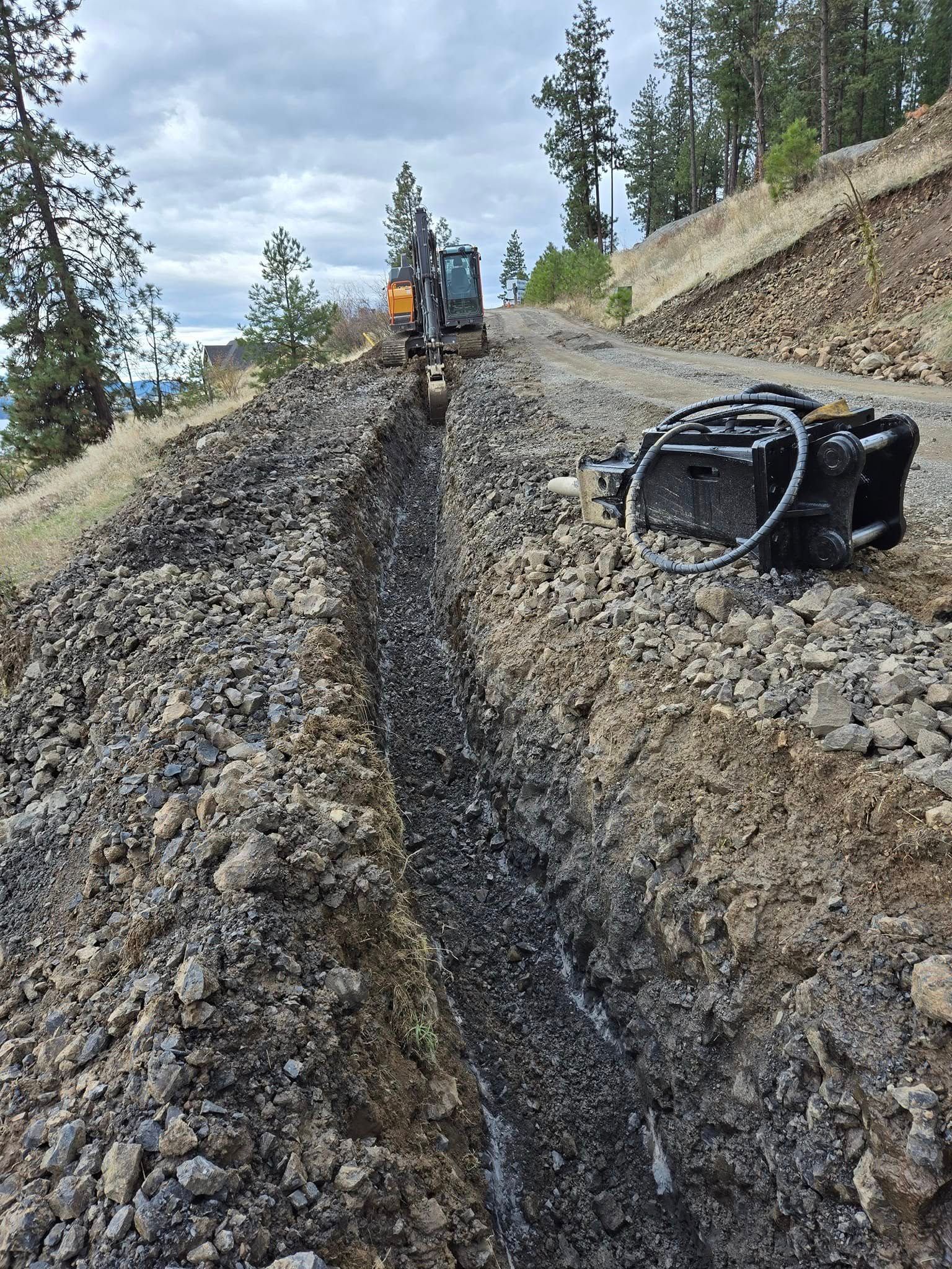 Excavator digging a trench on a hillside path for construction; cloudy sky.