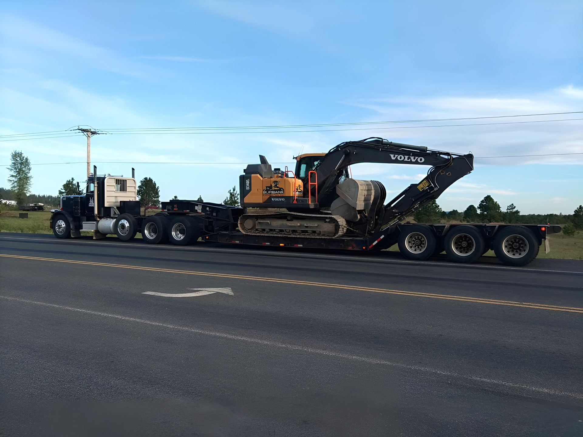 A semi-truck hauling a yellow and black excavator on a flatbed trailer on a road.