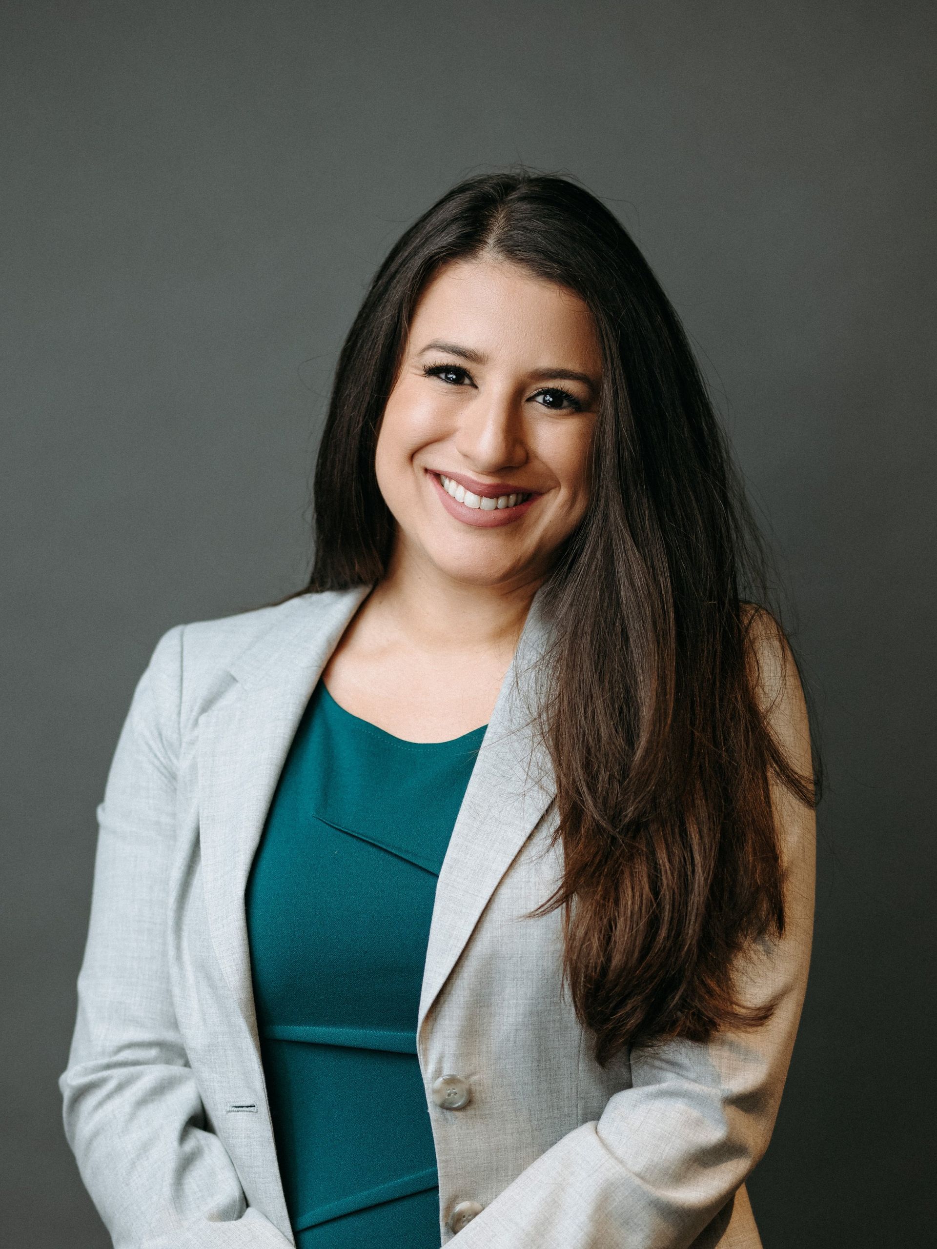 Woman with long dark hair smiles, wearing a gray blazer over a teal dress, against a gray backdrop.
