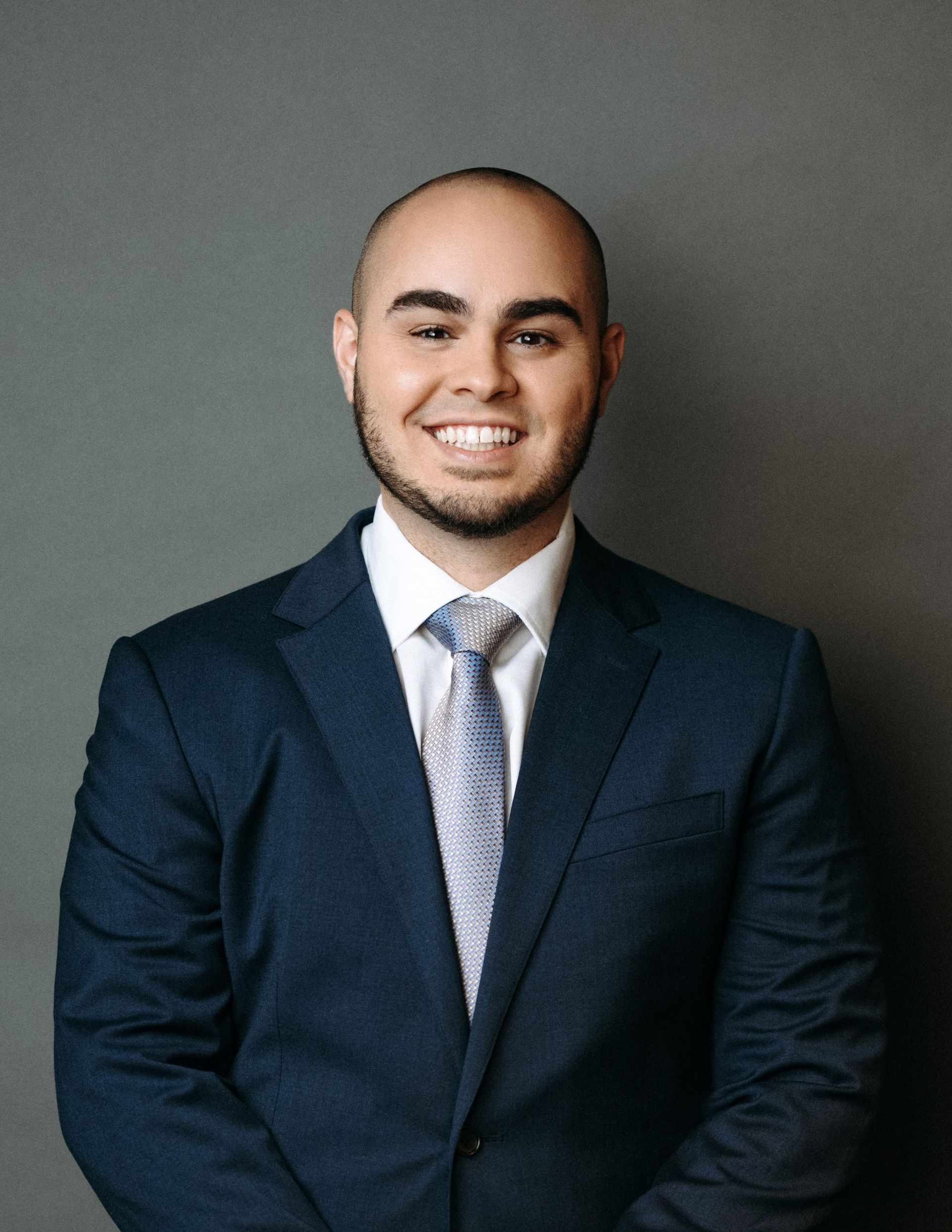 Man in a blue suit smiling, standing in front of a grey backdrop.